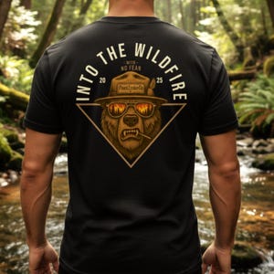 以下が含まれることがあります： サングラスと帽子をかぶったクマのグラフィックが特徴の黒いTシャツ。円形のデザインで「INTO THE WILDFIRE」の文字。クマはタバコをくわえており、「Pine Ridge Lumber Co.」の文字。
