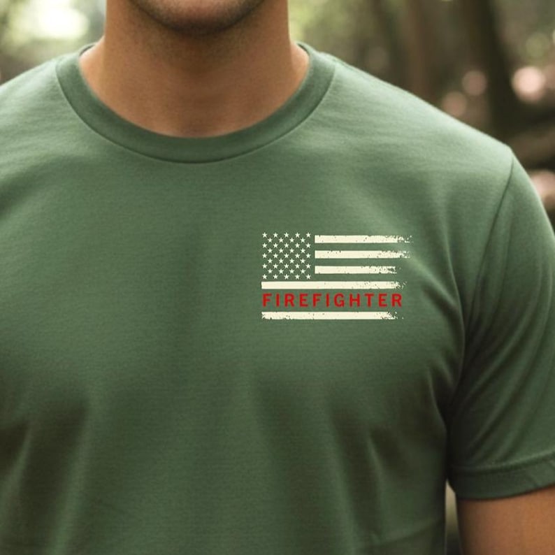 Close-up of a green T-shirt with a small distressed American flag graphic on the chest and a red stripe reading “Firefighter,” worn outdoors in a forest setting.