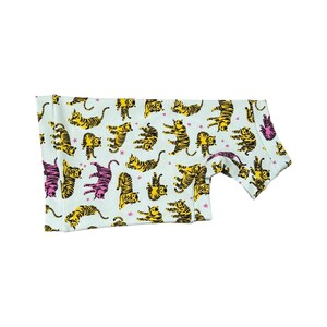 May include: A light blue garment featuring a repeating pattern of yellow and black tigers in various poses. Some tigers are colored in pink. The garment has a curved hemline and a short sleeve.