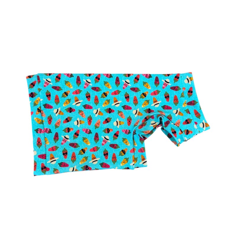 May include: A turquoise fabric with a repeating pattern of smiling chili peppers, watermelons, and other fruits. The fabric is folded, revealing a sleeve or edge, and is set against a white background.