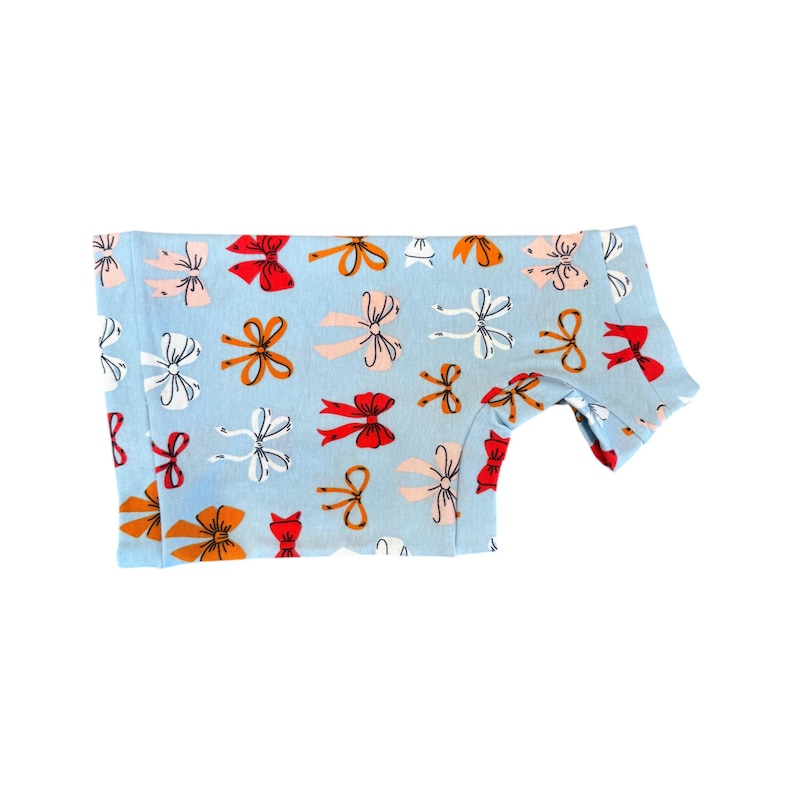 May include: Light blue dog shirt with a repeating pattern of colorful bows in red, orange, and white. The shirt is made of a soft material and has a short sleeve design.