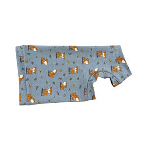 May include: A blue short-sleeved shirt with a repeating pattern of orange foxes and green leaves. The shirt is folded and appears to be made of a soft fabric. The foxes are in various poses.