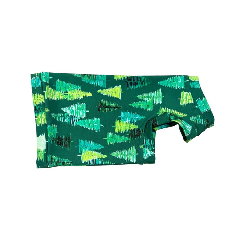 May include: Green fabric with a repeating pattern of stylized Christmas trees in shades of green and black. The fabric is folded, showing the pattern on both sides. The trees are drawn with a brushstroke effect.