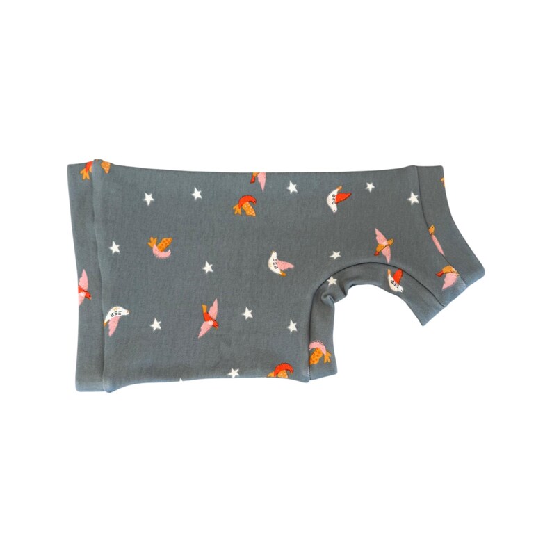 May include: A gray short-sleeved shirt with a pattern of pink, orange, and white birds and white stars. The shirt is folded and has a rounded neckline and short sleeves. The fabric appears to be soft and comfortable.