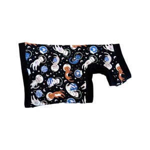 May include: Black onesie with a repeating pattern of cats in space suits, planets, and stars. The cats are in various colors, including white, orange, and blue. The onesie has black trim around the edges and sleeves.