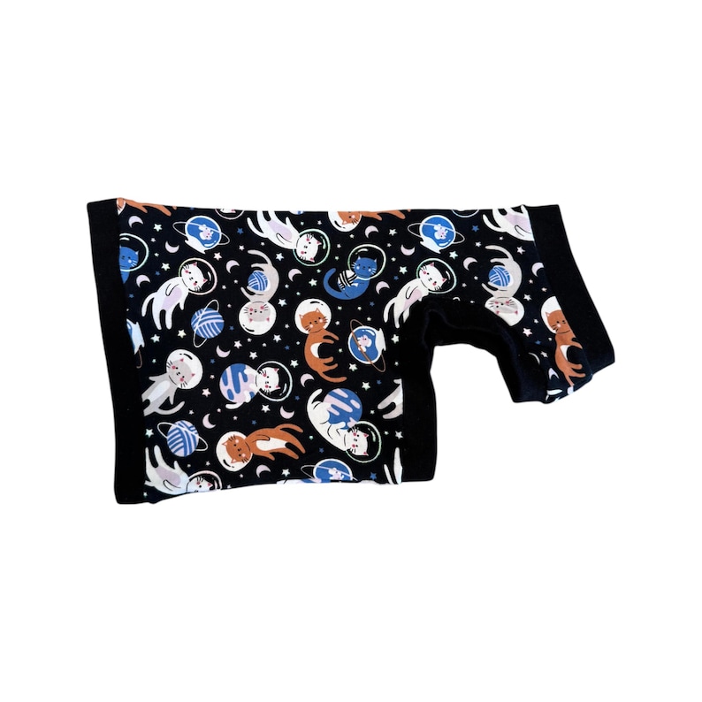 May include: Black onesie with a repeating pattern of cats in space suits, planets, and stars. The cats are in various colors, including white, orange, and blue. The onesie has black trim around the edges and sleeves.