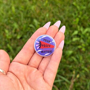 May include: A circular pinback button featuring a purple background with white abstract shapes and a red megaphone graphic. The text printed in black on the megaphone reads YOUR VOICE MATTERS, conveying a clear message in a compact design.
