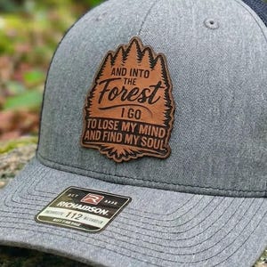 May include: Gray and navy blue trucker hat with a leather patch featuring the text "And Into The Forest I Go To Lose My Mind And Find My Soul." The hat has a curved brim and a mesh back.