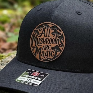 May include: Black trucker hat with a mesh back and a curved brim. The hat features a brown leather patch with the words "All Mushrooms Are Magic" in a circular design. The hat is sitting on a mossy rock.