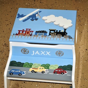 May include: A blue and white wooden step stool with a train, airplane, and cars painted on it. The top step has the name "Jaxx" painted on it.