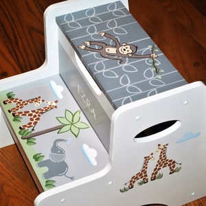 May include: White wooden two-step stool with a gray top featuring a monkey design. The bottom step has a giraffe, elephant, and palm tree design.