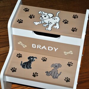 May include: A white and brown wooden step stool with a storage compartment. The stool is decorated with paw prints, bones, and cartoon dogs. The name "BRADY" is written on the front of the stool.