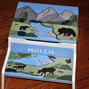 May include: A blue and white wooden step stool with a nature scene painted on the top and bottom steps. The top step features mountains, a river, trees, and bears. The bottom step features a river, trees, and deer. The step stool is personalized with the name "HOLLIS".