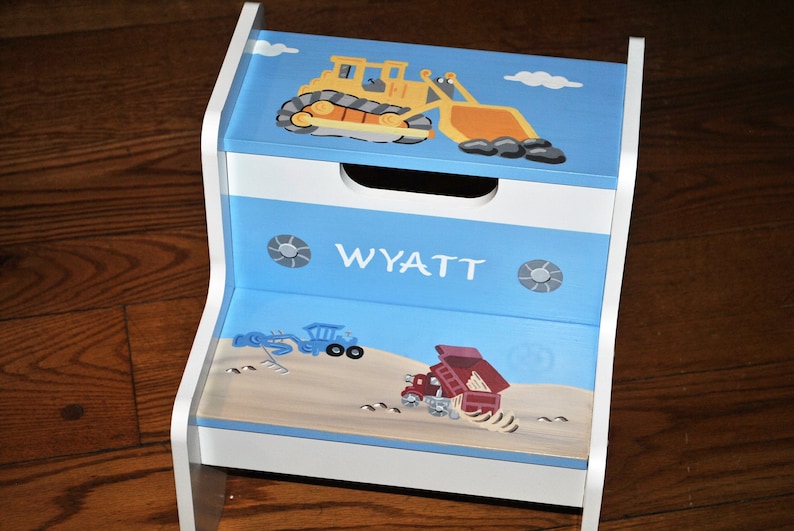 Pu&ograve; includere: Uno sgabello di legno blu e bianco con un veicolo da costruzione giallo e un camion ribaltabile rosso dipinti su di esso. Lo sgabello ha una parte superiore bianca con una striscia blu e il nome "WYATT" scritto su di esso.