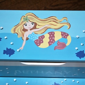 May include: A blue wooden step stool with a mermaid design. The mermaid has yellow hair, a pink and yellow tail, and is swimming in a blue ocean with white bubbles. The stool has a white drawer with the name "BETHANY" written on it.