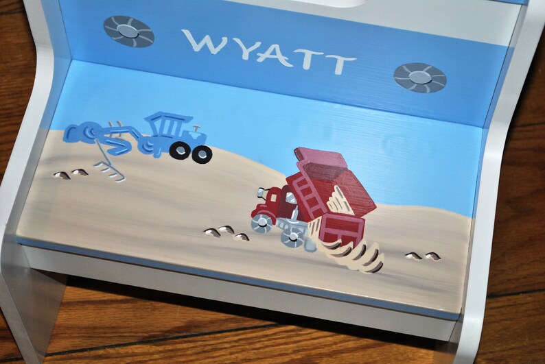 Pu&ograve; includere: Uno sgabello a gradini in legno blu e bianco con un piano superiore blu chiaro. Il piano superiore &egrave; dipinto con un camion ribaltabile rosso e un escavatore blu. Lo sgabello &egrave; personalizzato con il nome "WYATT" in lettere bianche.