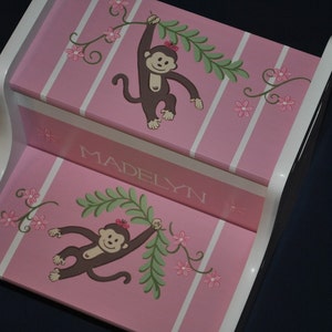 May include: Pink wooden step stool with white trim, featuring a monkey design with green leaves and pink flowers. The stool is personalized with the name "Madelyn".