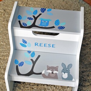 May include: A white wooden step stool with two steps. The steps are painted gray and decorated with blue tree branches, leaves, and an owl. The name "REESE" is written in blue. A fox and a rabbit are painted on the bottom step.