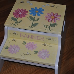 May include: A yellow wooden step stool with a white trim. The top and bottom steps are decorated with pink, blue, and yellow flowers and bees. The top step also has the name "Banner" written on it.