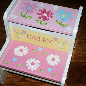 May include: A pink and yellow wooden step stool with a white trim. The top step has a pink surface with a blue, pink, and yellow flower design. The bottom step has a pink surface with white flowers and green leaves. The name "KAILEY" is written on the yellow surface of the step stool.