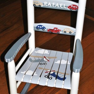 May include: White rocking chair with gray arms and a blue and white airplane design on the seat. The chair is painted with the name "Rafael" and has a red, blue, and yellow car design on the back.