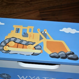 Pu&ograve; includere: Una scatola di legno blu per giocattoli con un bulldozer giallo dipinto sul coperchio. Il bulldozer trasporta pietre nel suo secchio. La scatola ha un cassetto bianco con il nome "WYATT" scritto su di esso.