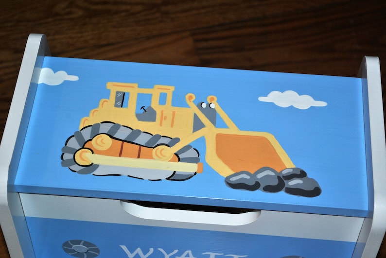 Pu&ograve; includere: Una scatola di legno blu per giocattoli con un bulldozer giallo dipinto sul coperchio. Il bulldozer trasporta pietre nel suo secchio. La scatola ha un cassetto bianco con il nome "WYATT" scritto su di esso.