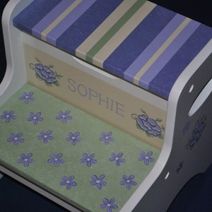 May include: White wooden step stool with a green and purple floral design and the name "Sophie" painted on the front. The top of the stool has a purple and green striped design.