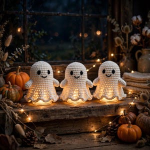 May include: Three white crocheted ghost figures with black embroidered eyes and mouths. Each ghost is illuminated from within by a small light, creating a soft glow. The figures are arranged on a wooden surface alongside several small orange pumpkins.