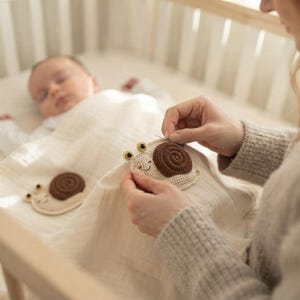 May include: A cream-colored muslin baby blanket featuring two crocheted snail appliqués. Each snail has a light beige body, a dark brown spiral shell, and two small eyes. The blanket is made of a soft, textured fabric with a visible weave pattern.
