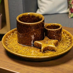 May include: A brown ceramic set with a star-shaped accent. The set includes a round plate with a textured pattern, two cylindrical cups, and a star-shaped piece. The pieces have a glossy finish and are displayed on a wooden surface.