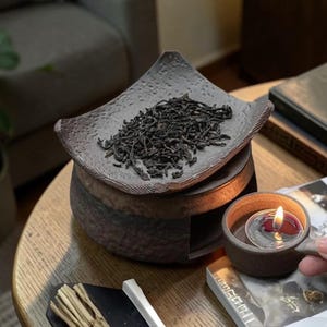May include: A dark brown ceramic tea warmer with a textured, curved top plate holding loose black tea leaves. A separate small matching ceramic cup with a handle contains a lit tea light candle, designed to be placed inside the warmer base.