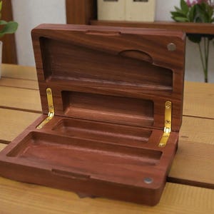 May include: Open wooden box with a hinged lid, revealing compartments. The box is made of dark brown wood with brass-coloured hinges. The interior features a small compartment and a larger rectangular space. The box is on a wooden surface.