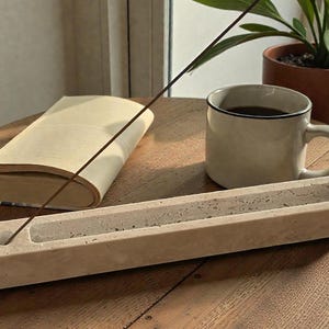 May include: A beige concrete incense holder with an incense stick, a mug of coffee, and a book on a wooden table. A potted plant is in the background. The incense holder is rectangular with a groove for the incense to sit in.