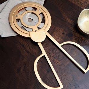 May include: A gold-colored, decorative kitchen tool with a circular design and scissor-like handles. The tool has a unique, geometric pattern and is placed on a dark wooden surface. A small, gold-colored scoop is also visible.