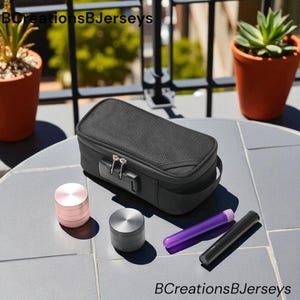 May include: Black zippered storage case with a combination lock, two metal grinders, and two tubes on a gray table. The case has a mesh top and a handle. The grinders are pink and silver. The tubes are purple and black. The text "BCreationsBJerseys" is visible.