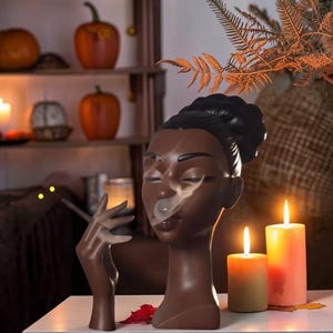 May include: A dark-skinned ceramic bust of a person with hair styled in a high bun, featuring a hand raised near the face holding a thin, dark stick. The sculpture has a smooth, matte finish and is positioned next to two lit pillar candles.