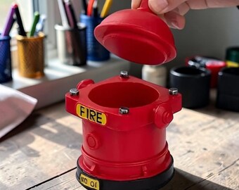 May include: A red fire hydrant-shaped container with a removable lid. The container has the word "FIRE" on a yellow label. The base is black, and the lid is being held open. The container is on a wooden surface.
