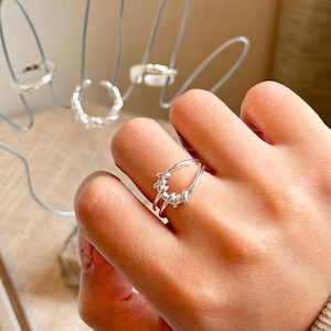 May include: A silver ring featuring a double-band design with a twisted, textured wire element that connects the two bands. The ring has a polished metallic finish and an open-ended structure, showcasing a continuous, flowing shape on the finger.