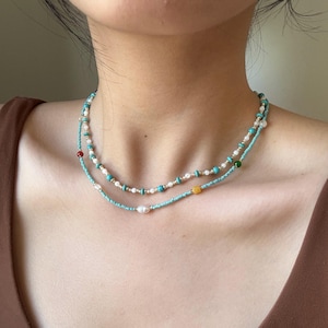 May include: A layered necklace set featuring two strands of small beads. The top strand consists of alternating turquoise and white beads. The bottom strand features turquoise beads, a single yellow square bead, and a central white pearl.