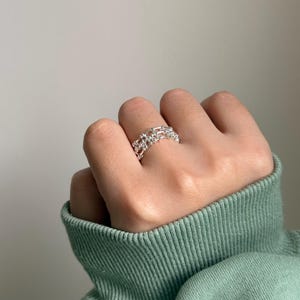 May include: A silver ring with a chain link design is worn on a finger. The ring has a double-layered band with multiple interconnected links. The background is a solid, neutral color, and the person is wearing a green sweater.