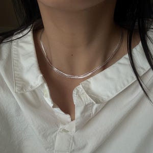 May include: A multi-strand silver-toned chain necklace featuring a delicate, woven texture. The necklace consists of several thin, interconnected strands that drape closely around the neck, creating a layered appearance against a white collared shirt.