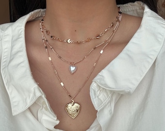 May include: A layered gold-tone necklace set featuring three distinct chains. The top chain has small heart-shaped links, the middle chain holds a white heart-shaped pendant, and the bottom chain features a large, ornate heart-shaped locket.