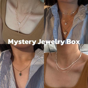 May include: A collection of four gold-toned necklaces featuring various designs, including layered chains, a heart-shaped pendant, a green oval stone pendant, and beaded strands. The image includes the text Mystery Jewelry Box.