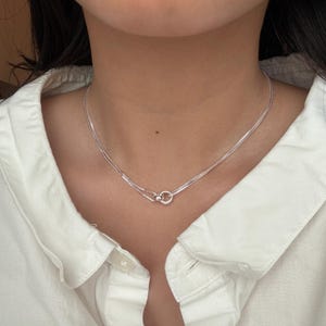Peut inclure: Un délicat collier en argent avec un design à double chaîne. Le collier présente un petit pendentif circulaire avec une perle de connexion. Le bijou est présenté sur une chemise blanche.