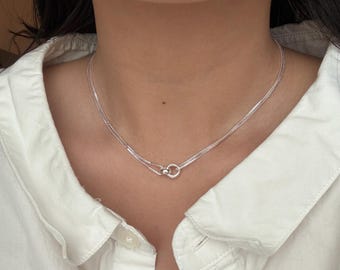 May include: A silver-toned necklace featuring two delicate, thin chains that meet at a small, circular ring clasp positioned at the center of the collarbone. The metallic chains have a fine, reflective texture and rest against the neck.