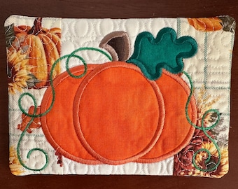 May include: A quilted rectangular placemat featuring an orange pumpkin with green leaves and vines. The pumpkin is the focal point, with brown stem and detailed stitching. The corners display fall-themed fabric with pumpkins and floral patterns.