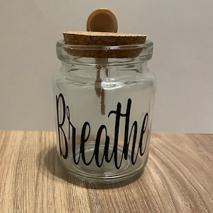 May include: A clear glass jar with a cork lid and a wooden spoon. The jar has the word "Breathe" written on it in black.