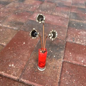 May include: A small bouquet of three silver flowers made from shotgun shells, arranged in a red shotgun shell vase.