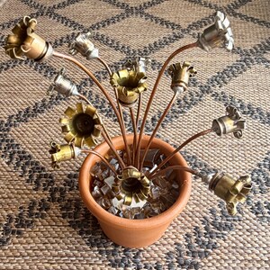 May include: A decorative arrangement featuring multiple metallic flower-shaped components made from brass and silver-toned casings. These are attached to thin copper-colored stems, all anchored within a small terracotta pot filled with clear glass.